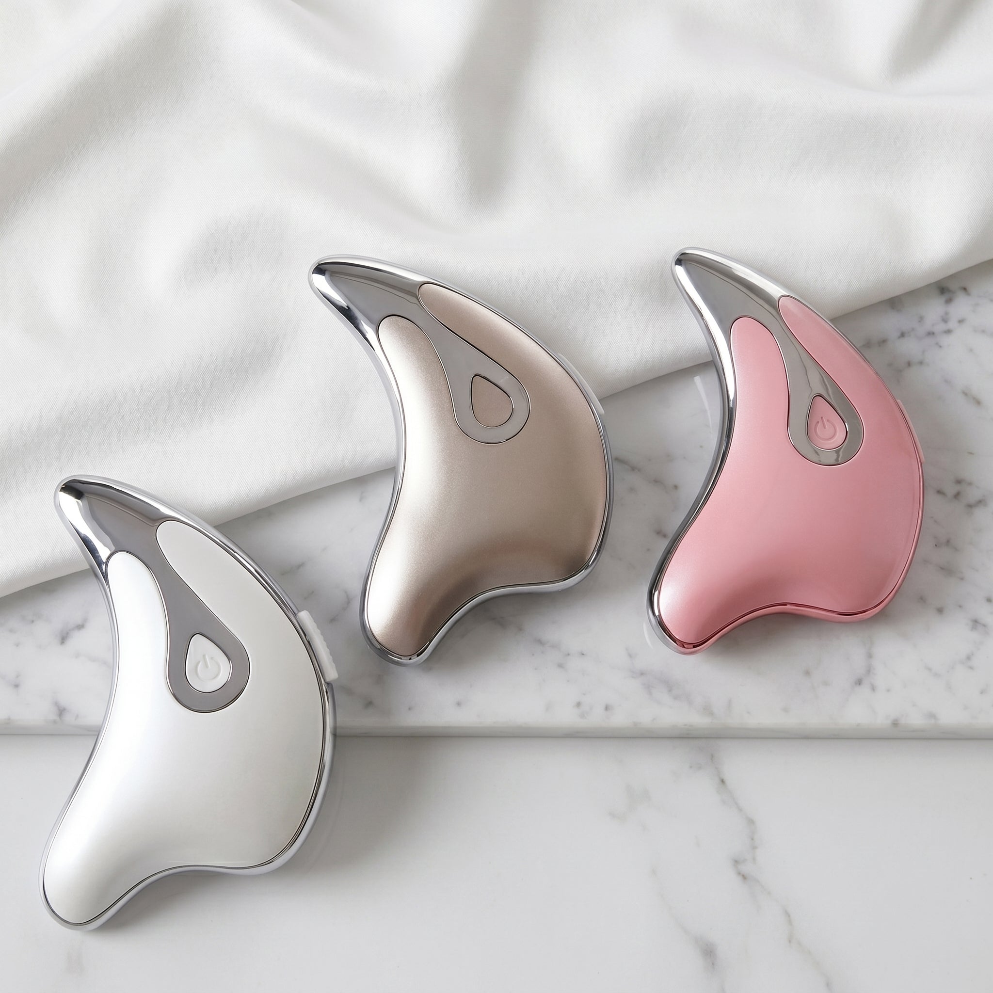 Three gua sha tools in silver, pink, and white.